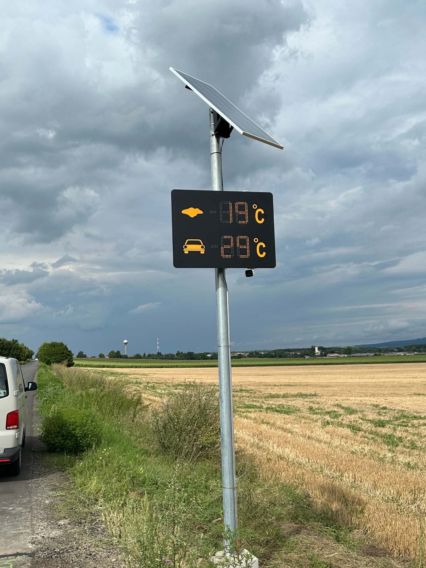 Cestná meteo tabuľa BELLIMPEX zobrazujúca teplotu vzduchu a vozovky pod dramatickou oblohou.